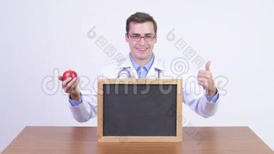 年轻快乐的帅哥医生拿着<strong>苹果</strong>对着木桌<strong>展示</strong>黑板